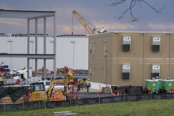 Yellow construction equipment, such as a crane, and gray metal beams are at a construction site at a data center campus near Adamstown, Maryland. The photo also shows two buildings and three workers wearing fluorescent vests.