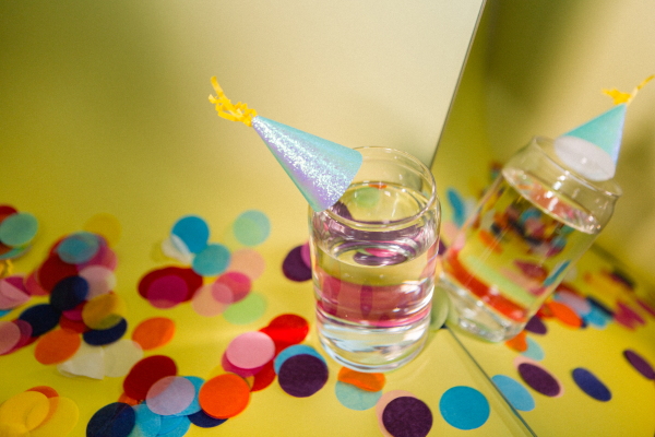 Photograph showing a glass of water looking at itself in the mirror against a yellow backdrop. The water glass is wearing a miniature party hat and is surrounded by confetti on the ground.