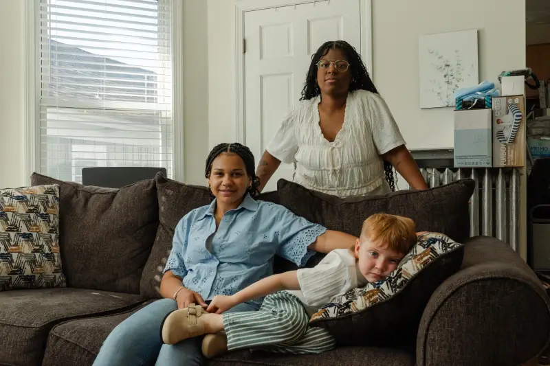 Emani Pike, 14, is sitting on a couch with Austin Pike, 4, who is laying down. Marissa stand behind them. Emani has long, dark braids and is wearing a blue button down shirt with jeans. Austin has red hair and is wearing a white shirt with white and green striped pants.