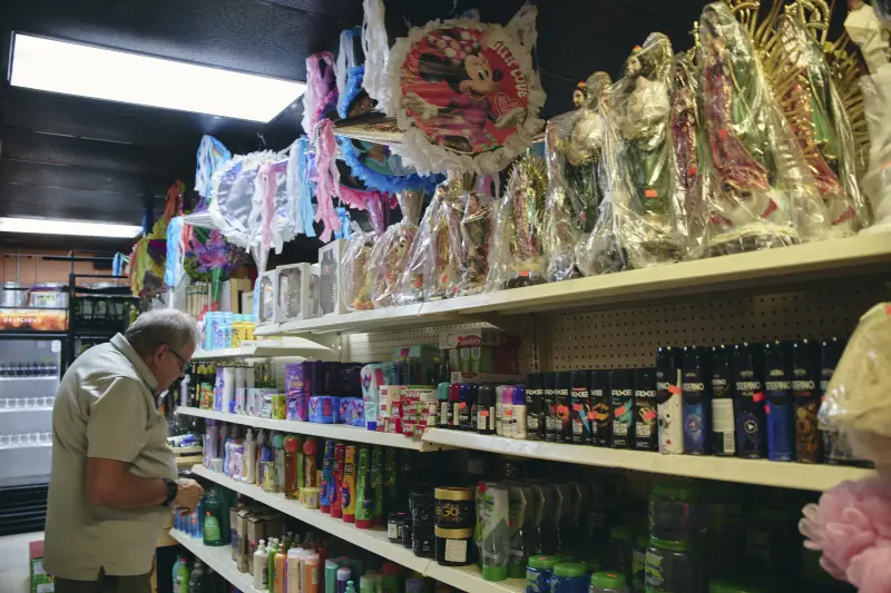 A man stands in front of store shelves inside La Guerrerense, a family-owned shop. The shelves have rows of grooming products, and a top shelf has a row of religious statues.