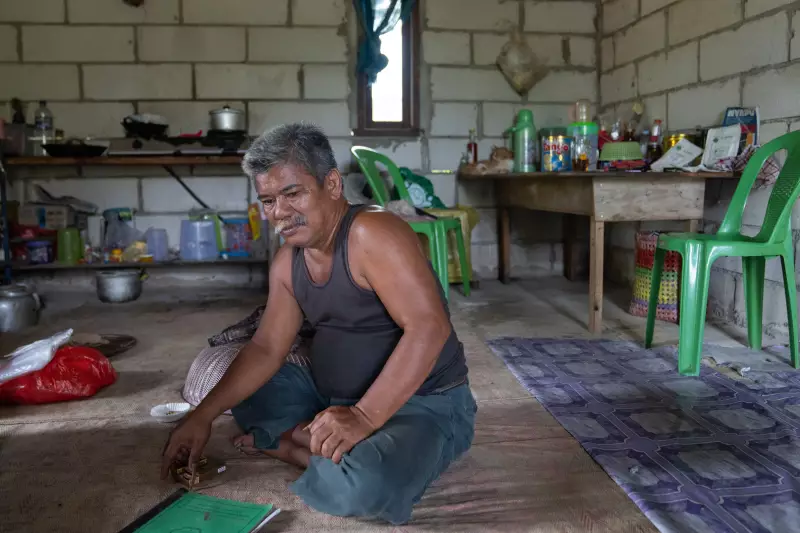 Pandi, 53, sits cross-legged on the floor with a bound document on the floor in front of him. Behind him are a gas stove, a table with kitchen items on it and two green plastic chairs. The walls behind him are composed of concrete blocks.