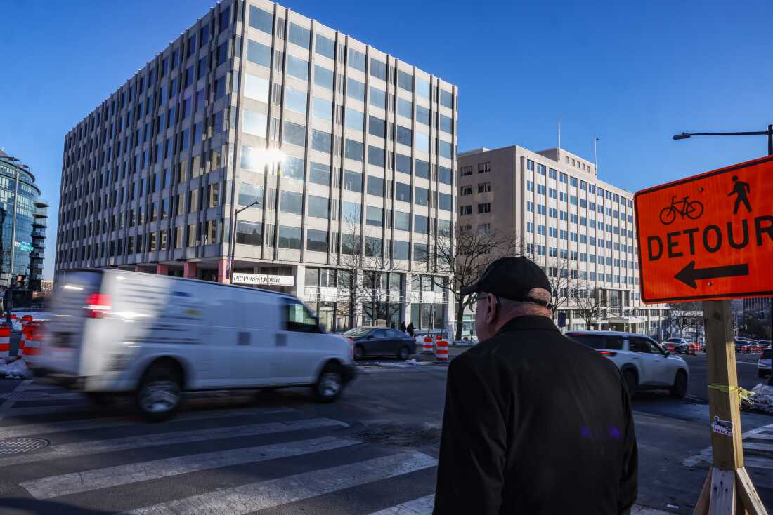 A concrete and glass building is seen across the street as an older man waits at a busy crosswalk.