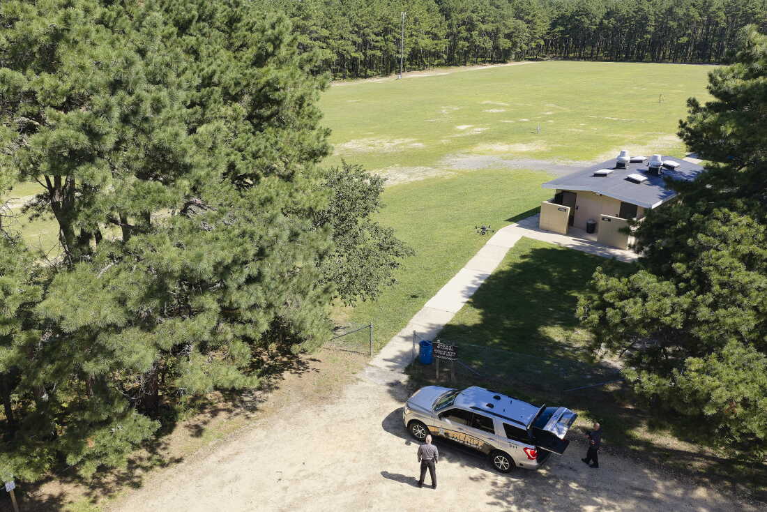 An aerial view of a drone as it lifts off near an open area that has trees around the edges. Two men stand near an SUV.