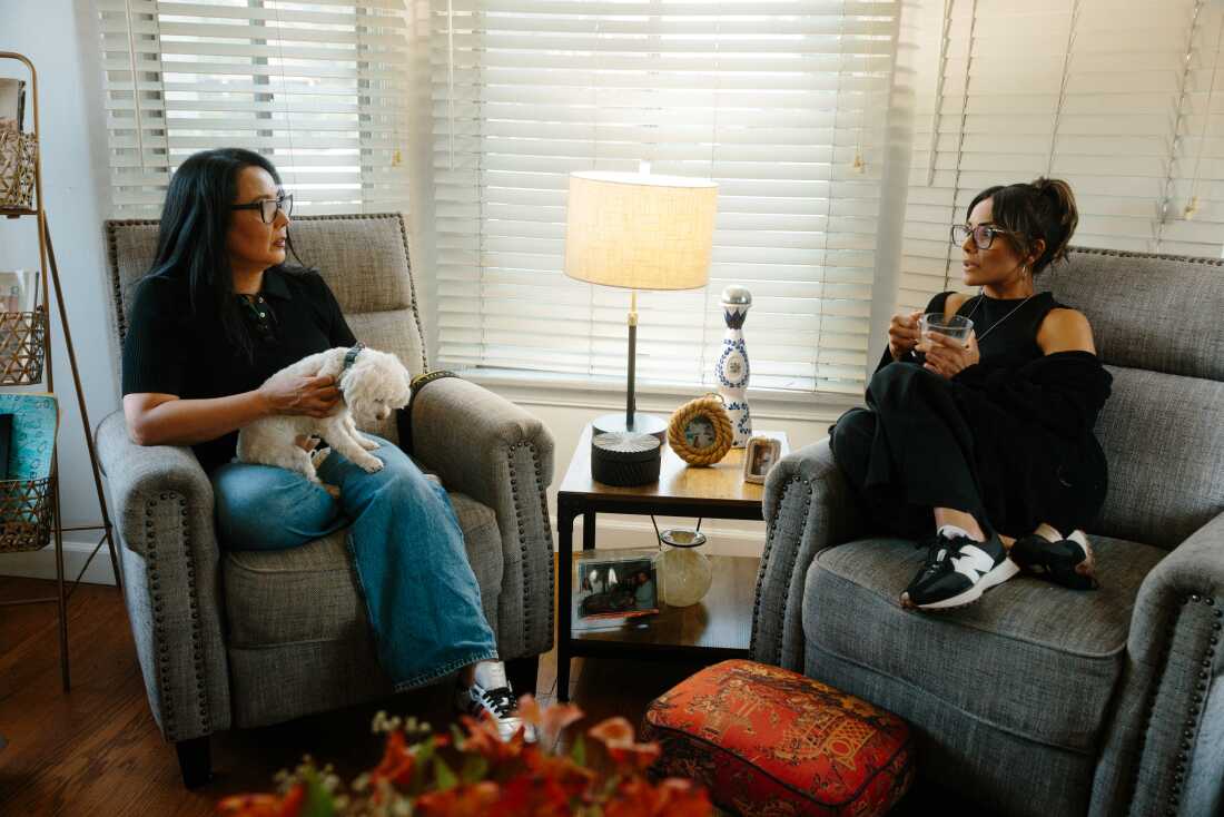 Dessaint (left) and Serrato (right) catch up on news from the Middle East. They are sitting in gray chairs in the living room. Dessaint holds a small, white dog on her lap.
