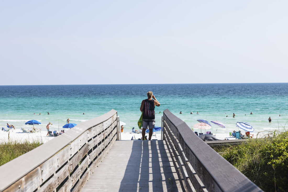 A man wearing shorts and a sleeveless top stands on the wooden boardwalk that leads to the sand at Ed Walline Regional Beach Access in Santa Rosa Beach, Florida, on August 25. Blue waters are in the background.