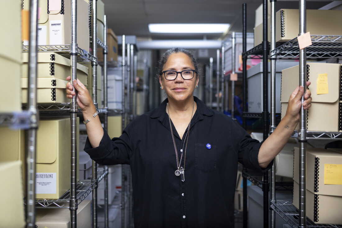 Laurice Bell stands between two rows of metal shelves holding boxes of archival materials that document the history of Black people in Evanston, Illinois.