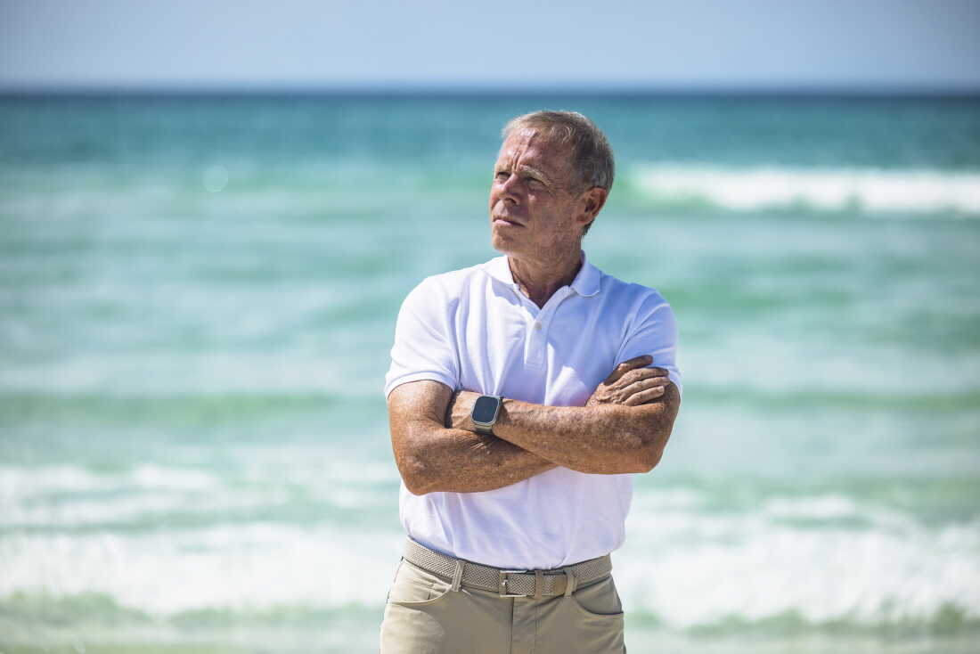 Dave Rauschkolb stands on a beach off Scenic Highway 30A on August 20. He's wearing a white polo shirt and khaki pants, and his arms are crossed across his chest. Water and waves are behind him.