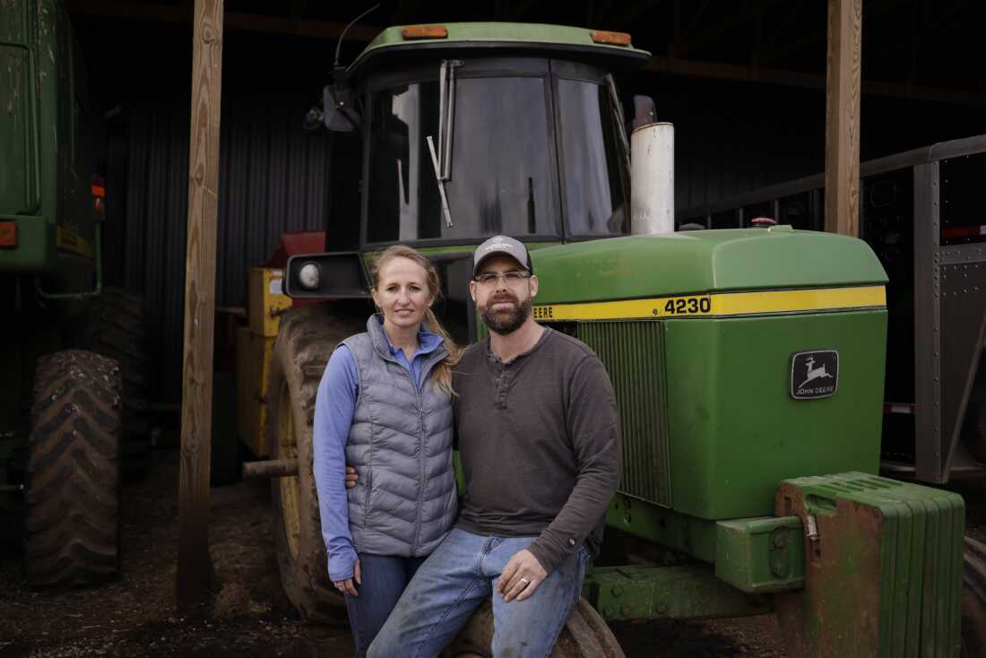 Amanda Hall stands next to a green tractor, and Reid Hall sits on the tractor's front wheel, with his arm around Amanda Hall's waist.