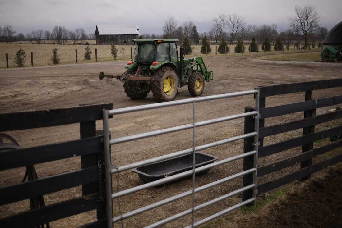 A person drives a John Deere tractor down a dirt road at Hallstead Farms. A wooden fence with a metal gate is in the foreground. A wooden barn is in the background.