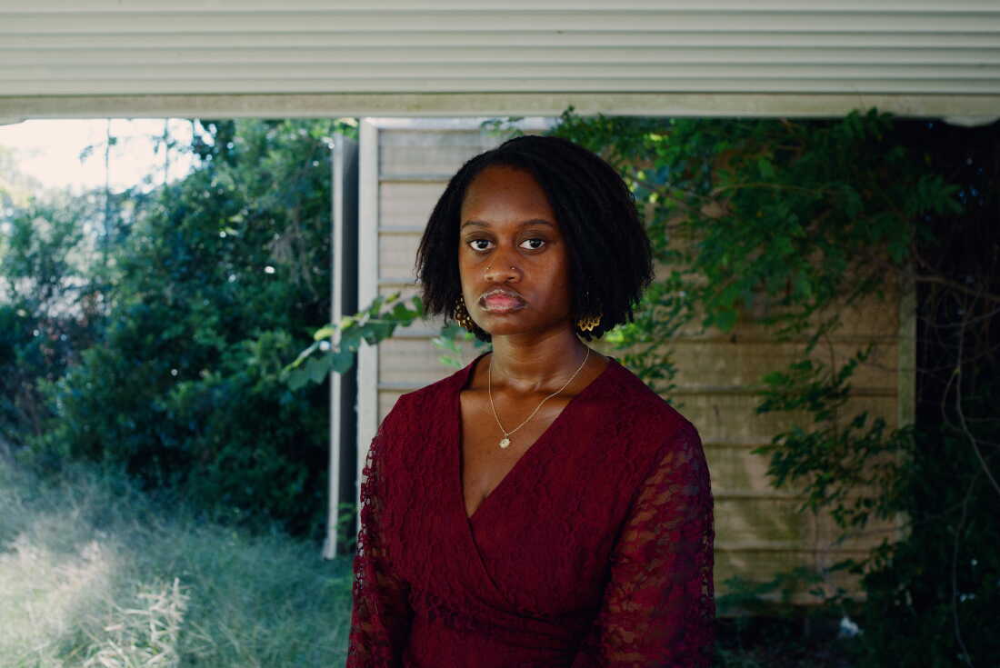 JoJo Kimble has chin-length dark, curly hair and is wearing a burgundy top with lace sleeves. She is standing under an awning with greenery behind her.