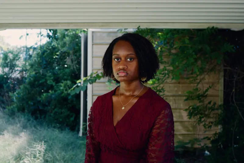 JoJo Kimble has chin-length dark, curly hair and is wearing a burgundy top with lace sleeves. She is standing under an awning with greenery behind her.