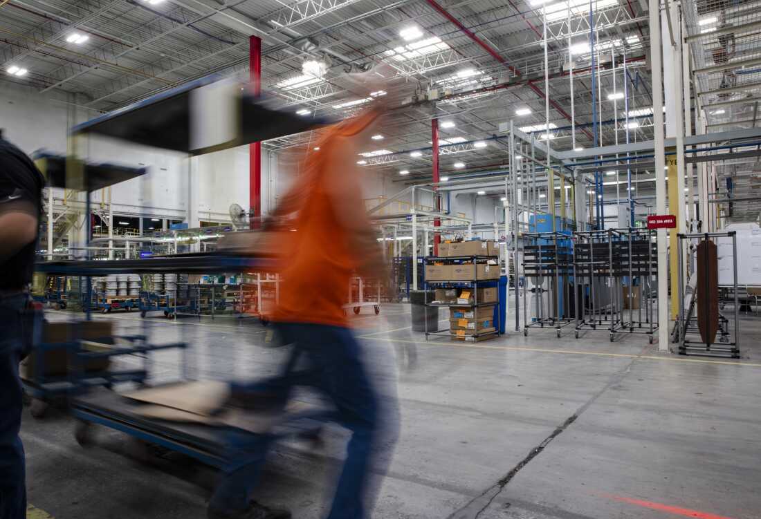 A worker, wearing an orange vest and photographed as a moving blur, pulls a cart behind them as the worker walks across the factory floor.