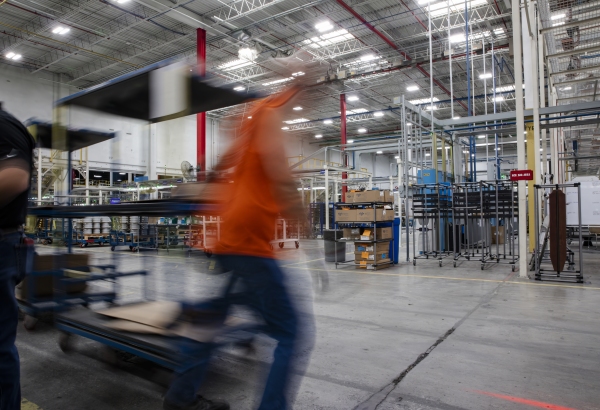 A worker, wearing an orange vest and photographed as a moving blur, pulls a cart behind them as the worker walks across the factory floor.