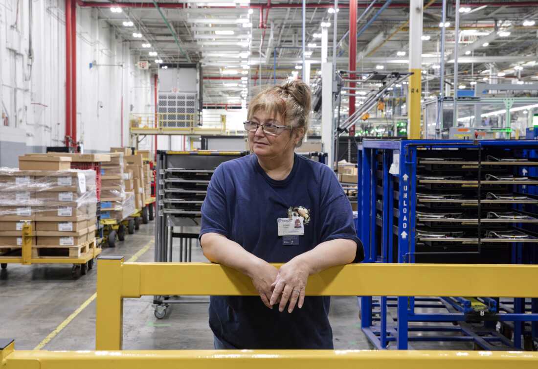 Doris Hamby stands on the factory floor. She's wearing a navy blue T-shirt and glasses.