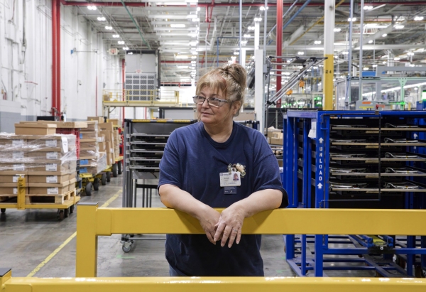 Doris Hamby stands on the factory floor. She's wearing a navy blue T-shirt and glasses.