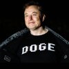 In this photo, Elon Musk is wearing a black shirt with "DOGE" printed on the front in large white capital letters. He's using his hands to hold wide open the dark jacket he's wearing over the shirt. Musk is smiling, and the background behind him is black.