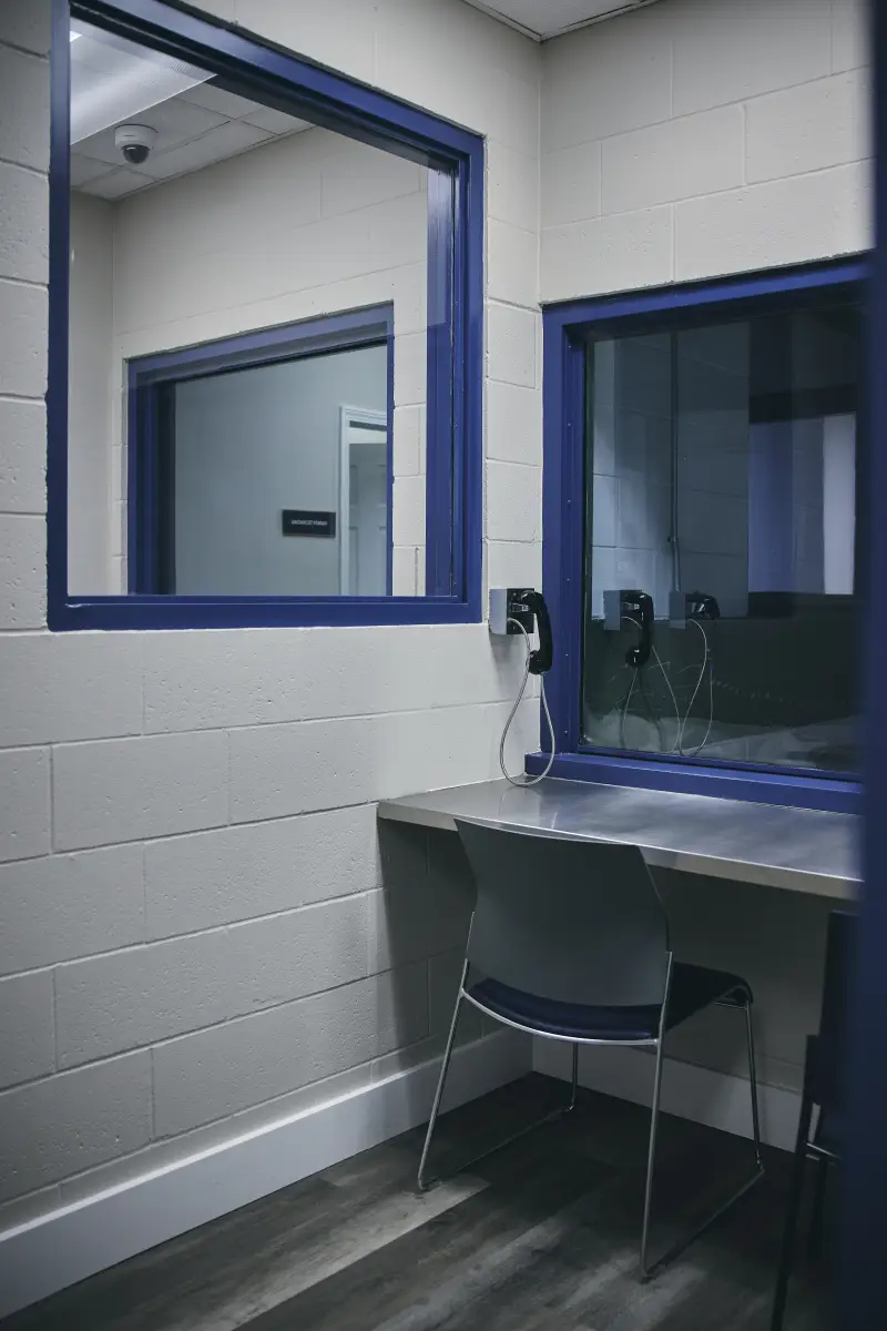 This visitation room at the Walton County Jail has a chair in front of an interior window and a phone on the wall, next to the chair.