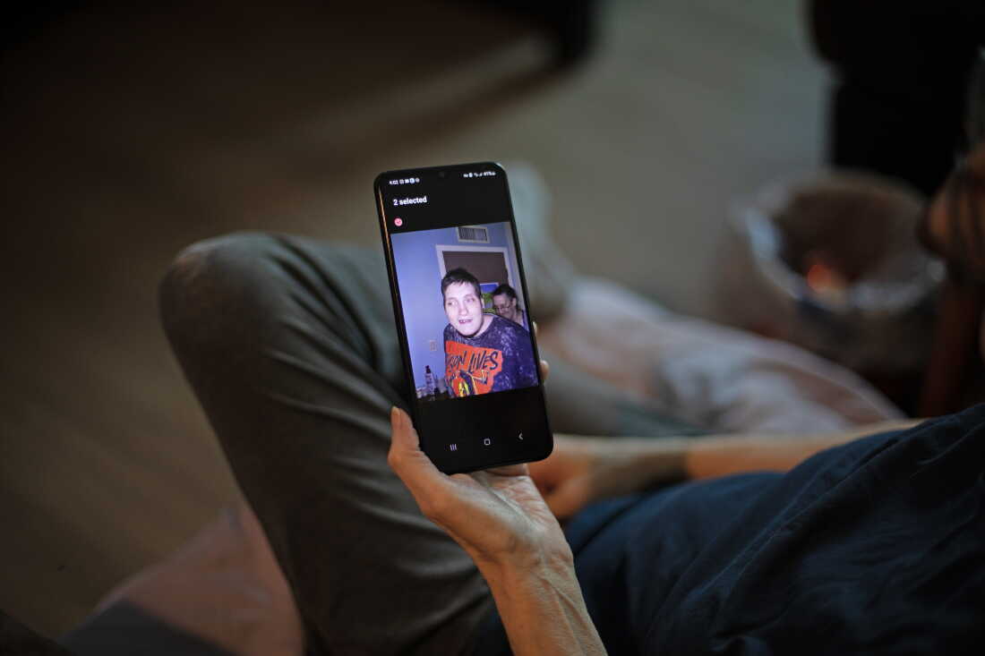 Debbie Willson holds her smartphone, which is displaying a photo of her son, Adam Willson, who died at age 27 in 2024. Adam is wearing a T-shirt that says "Jason Lives," with an image of the horror movie character Jason holding an axe above his head.