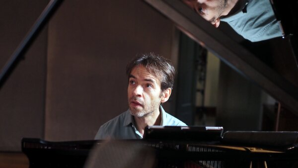 Vadim Neselovskyi looks off to the side while he sits at a piano. He wars a light blue collared shirt. His reflection is on the hood of the piano.