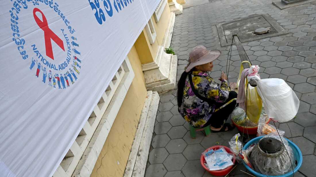 A vendor sits next to a logo for World AIDS Day, along a sidewalk in Phnom Penh on December 1, 2022. (Photo by TANG CHHIN Sothy / AFP) (Photo by TANG CHHIN SOTHY/AFP via Getty Images)