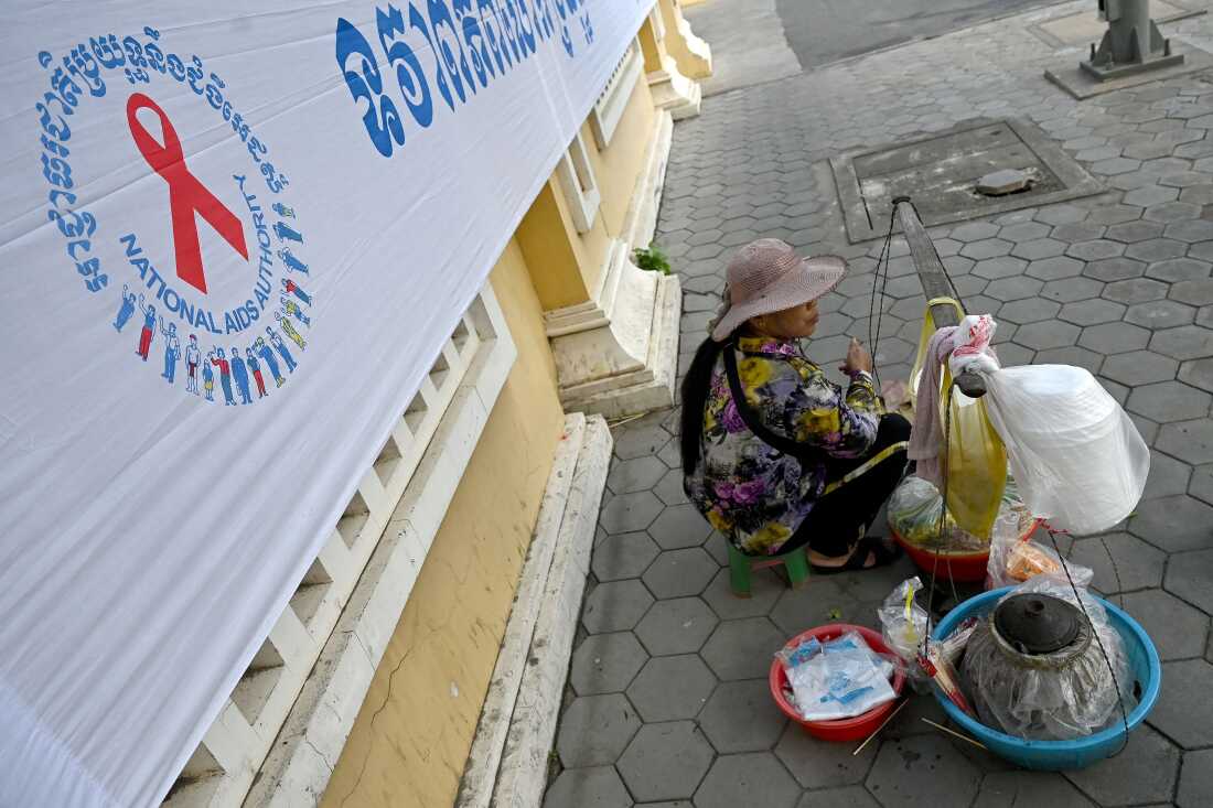 A vendor sits next to a logo for World AIDS Day, along a sidewalk in Phnom Penh on December 1, 2022. (Photo by TANG CHHIN Sothy / AFP) (Photo by TANG CHHIN SOTHY/AFP via Getty Images)