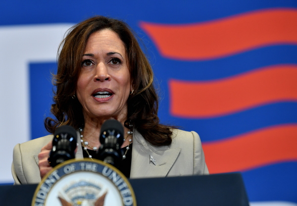 A close up of Vice President Kamala Harris. She is at a podium in front of a huge red and blue "Biden Harris" poster. She is wearing a beige suit and a pearl and gold necklace.