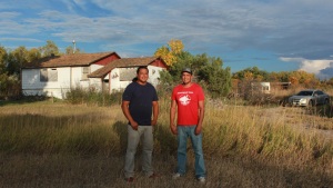 Lonny and Teyon Fritzler stand outside their childhood home. Plywood covers the windows of the white house, and tall gold-colored grass is high in the yard between the brothers and the house.
