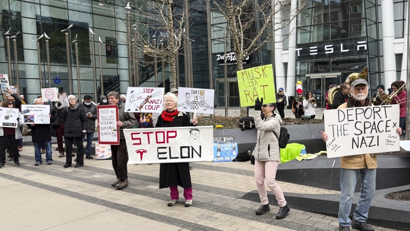 Protesters holding signs rally outside a Tesla store in Boston. One sign says "Stop Elon." 