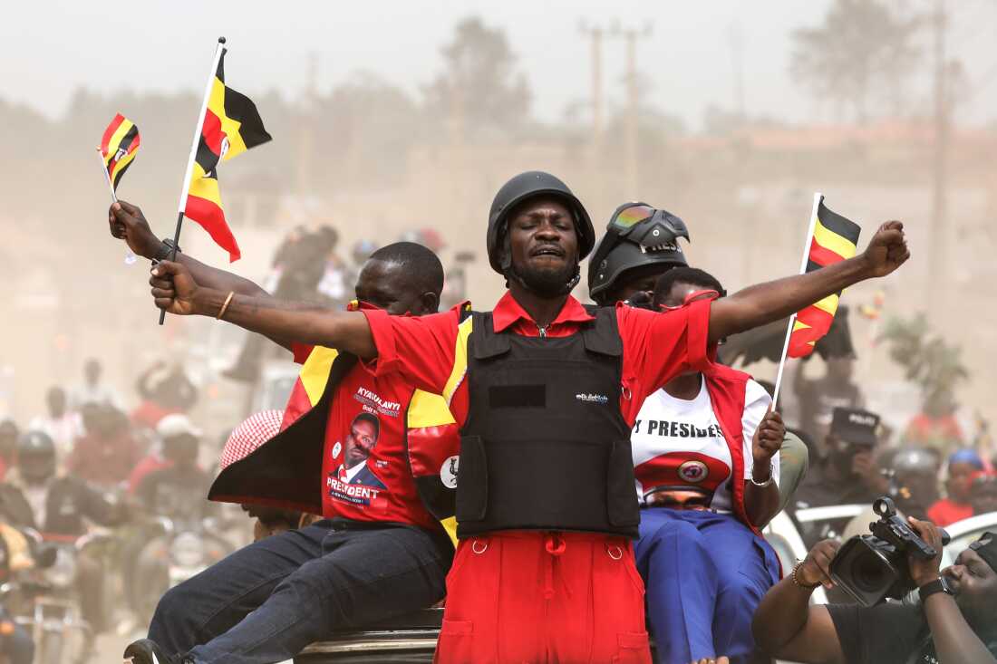 Ugandan opposition presidential candidate Bobi Wine waves to supporters at an election campaign rally in Mukono, Uganda, on January 9. He is wearing a short-sleeved red top, a black flak jacket and a black helmet, while holding a small Ugandan flag in one hand.