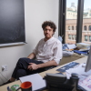 Tristan Buckmaster, mathematician at New York University, poses for a portrait in his office in Manhattan. He is wearing a white button down shirt and dark slacks. Behind him is a blackboard and a window. In front of him is a desk with a computer, keyboard and some papers.
