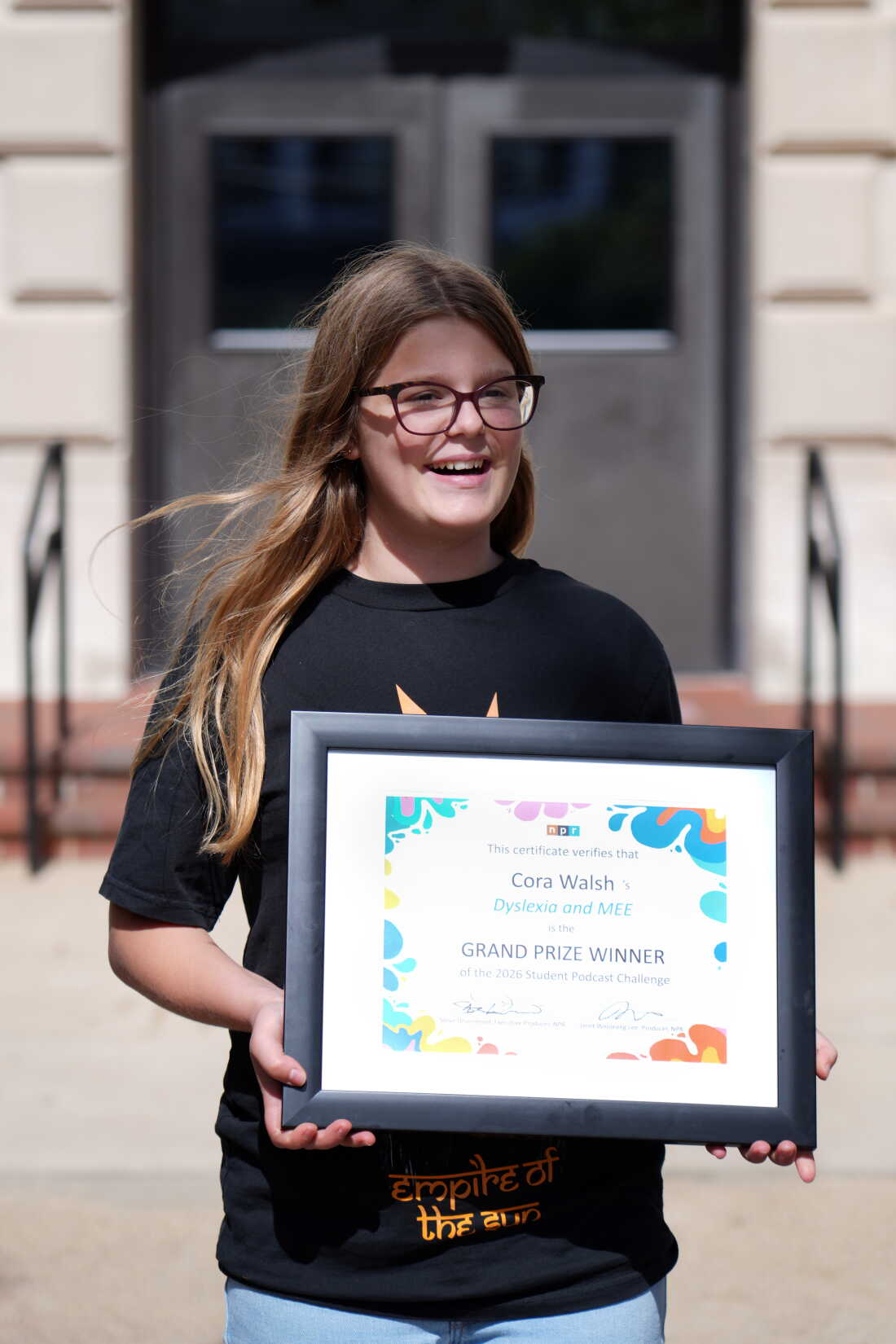 Cora Walsh stands outdoors holding a framed certificate declaring her the grand prize winner of the 2026 NPR Student Podcast Challenge.