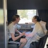 Peter and Aubrie in their accessible room on the California Zephyr Amtrak train. They sit next to a window facing each other. Aubrie’s wheelchair is to her side. Outside the window, water and trees rush by.