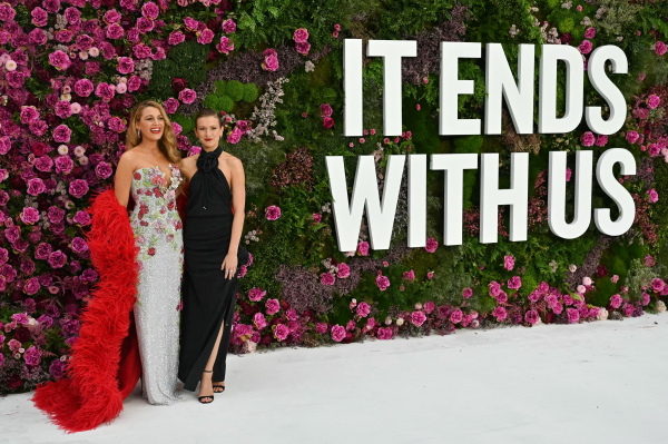 This photo shows two actresses from the movie "It Ends With Us" dressed in formal gowns and standing against a backdrop that's made up of fuchsia-colored flowers and green foliage. The backdrop says in big white letters "It Ends With Us."