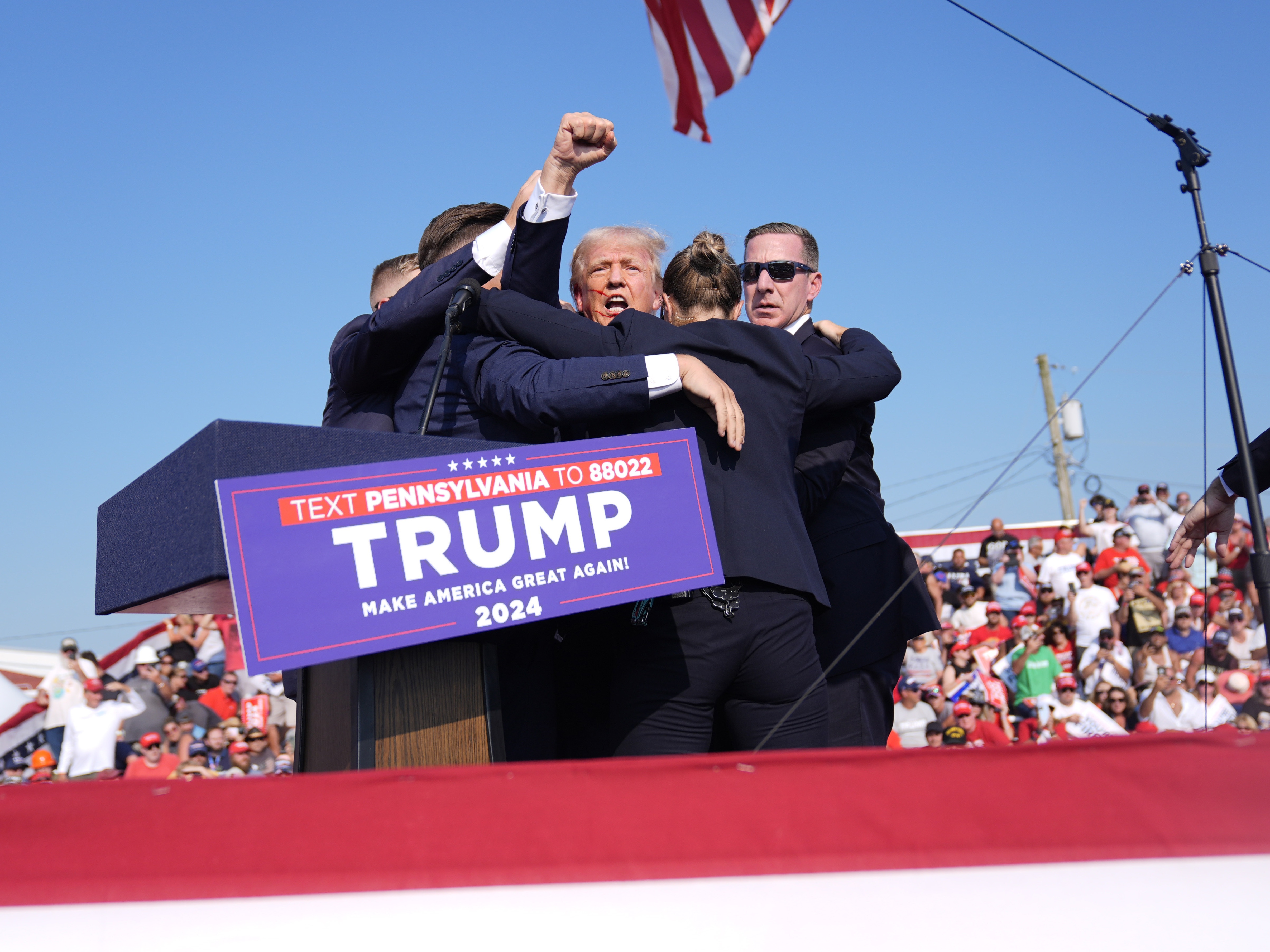 Trump is holding a fist in the air as Secret Service agents surround him as he leaves the stage at the campaign rally in Butler on July 13. The agents are wearing dark blue suits. Behind the stage are rally attendees.