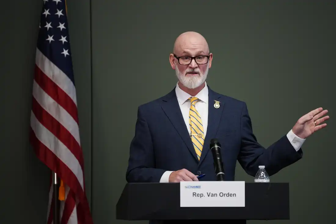Representative Derrick Van Orden speaks in Eau Claire, Wisconsin, on October 20. He is wearing a suit and is standing behind a lectern with a microphone attached to it. On the left side of the frame is a U.S. flag on a flagpole.