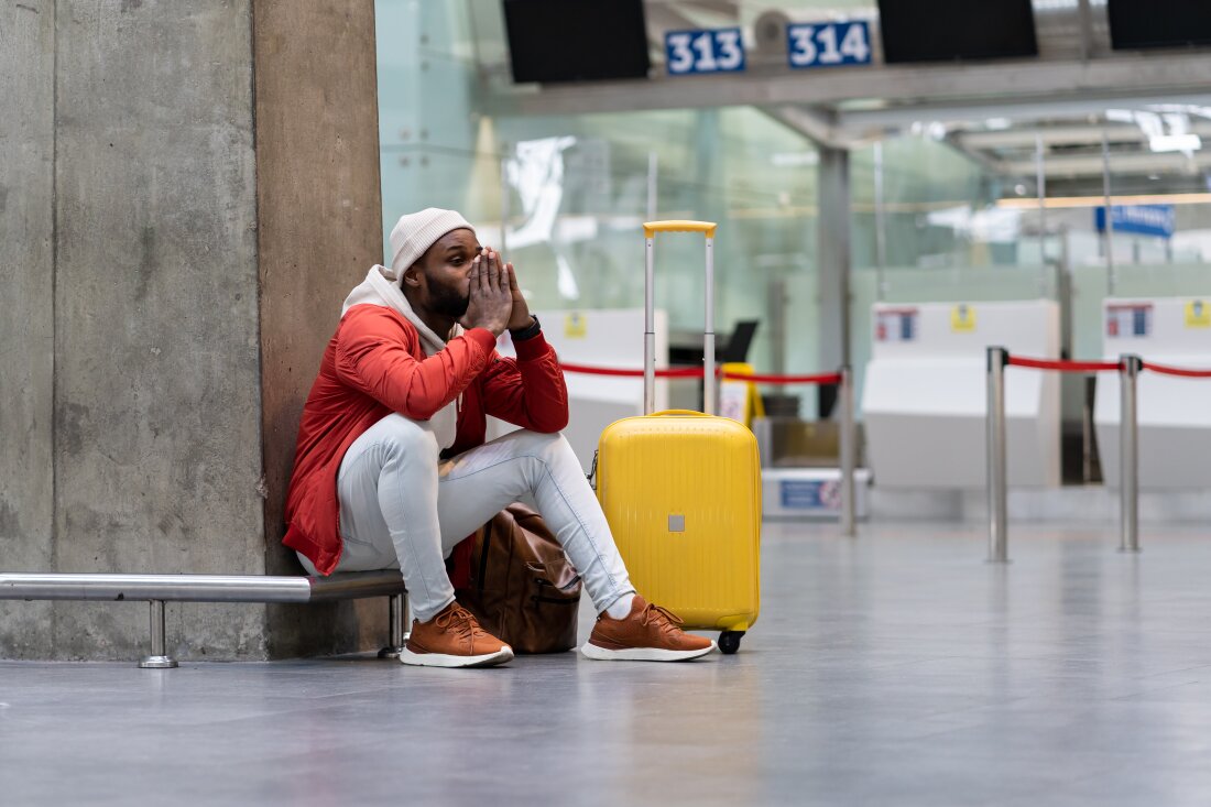 A young Black man sits on the ground inside an airport next to his yellow rolling suitcase, waiting to check in for his flight. He wears a red puffer coat and a toboggan and puts his head in his hands in frustration over travel delays.