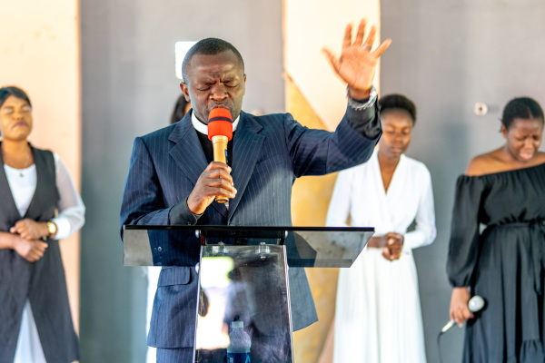 In this photo, Pastor Billy is standing behind a lectern and holding a microphone as he delivers a sermon to his congregants at the Somone Community Center. He's wearing a dark blue suit, and behind him stand four people, at least three with their eyes closed.