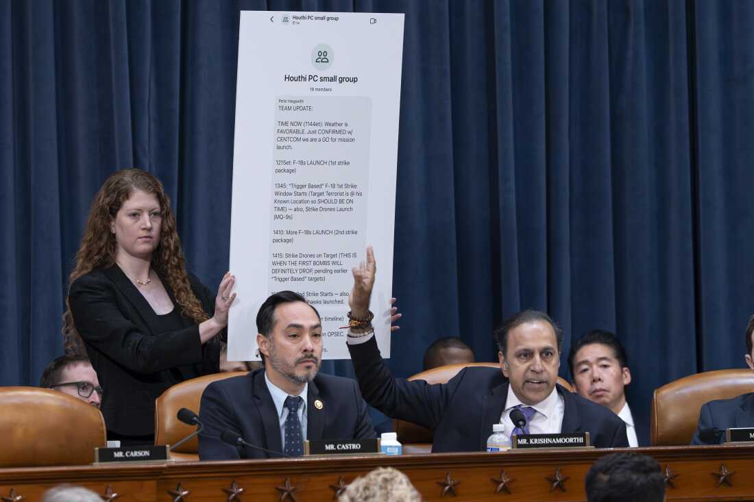In this photo, Rep. Raja Krishnamoorthi, seated at a committee hearing, points to a large chart behind him that displays an enlarged text message from a group chat. A woman is holding up the chart, and Rep. Joaquin Castro is seated next to Krishnamoorthi.