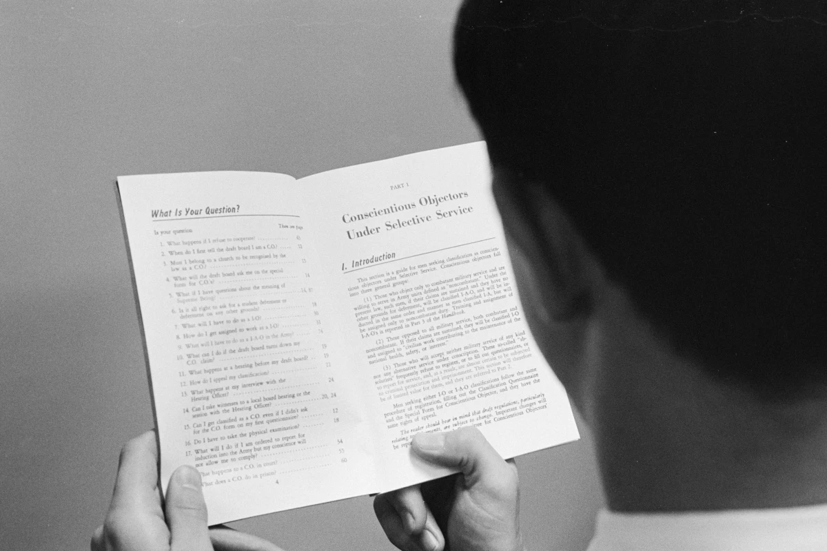 In 1965, a man looks over a booklet that's opened to a page with the heading 'Conscientious Objectors Under Selective Service.'