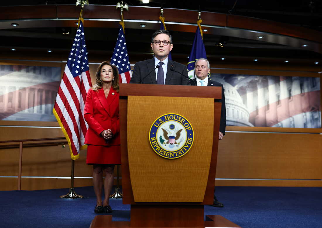 House Speaker Mike Johnson stands behind a lectern with microphones attached to it as he addresses reporters during a news conference Tuesday. Representative Lisa McClain and Majority Leader Steve Scalise stand behind Johnson.