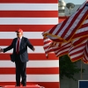 Wearing a suit and red baseball cap, President Trump stands onstage with arms outstretched to deliver remarks at the Iowa State Fairgrounds on July 3, 2025. Multiple American flags wave on flagpoles on the right side of the frame.