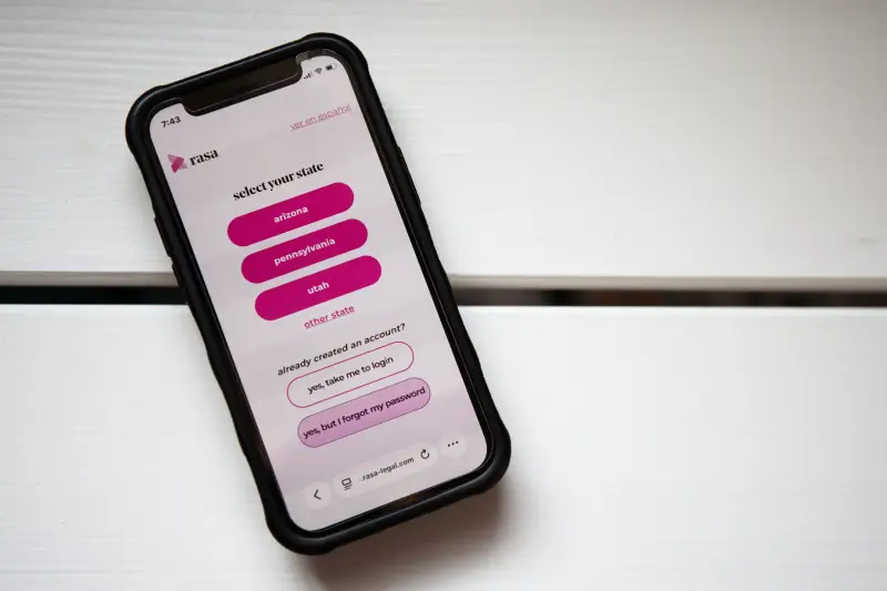 A photo of a smartphone showing the Rasa Legal website, which is a white screen with pink buttons. The screen says "select your state" and gives Arizona, Pennsylvania and Utah as options, along with "other state."