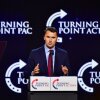 In this photo, Charlie Kirk stands at a lectern and speaks into a microphone attached to it. He's wearing a suit, and behind him is a backdrop that says "Turning Point PAC" multiple times.