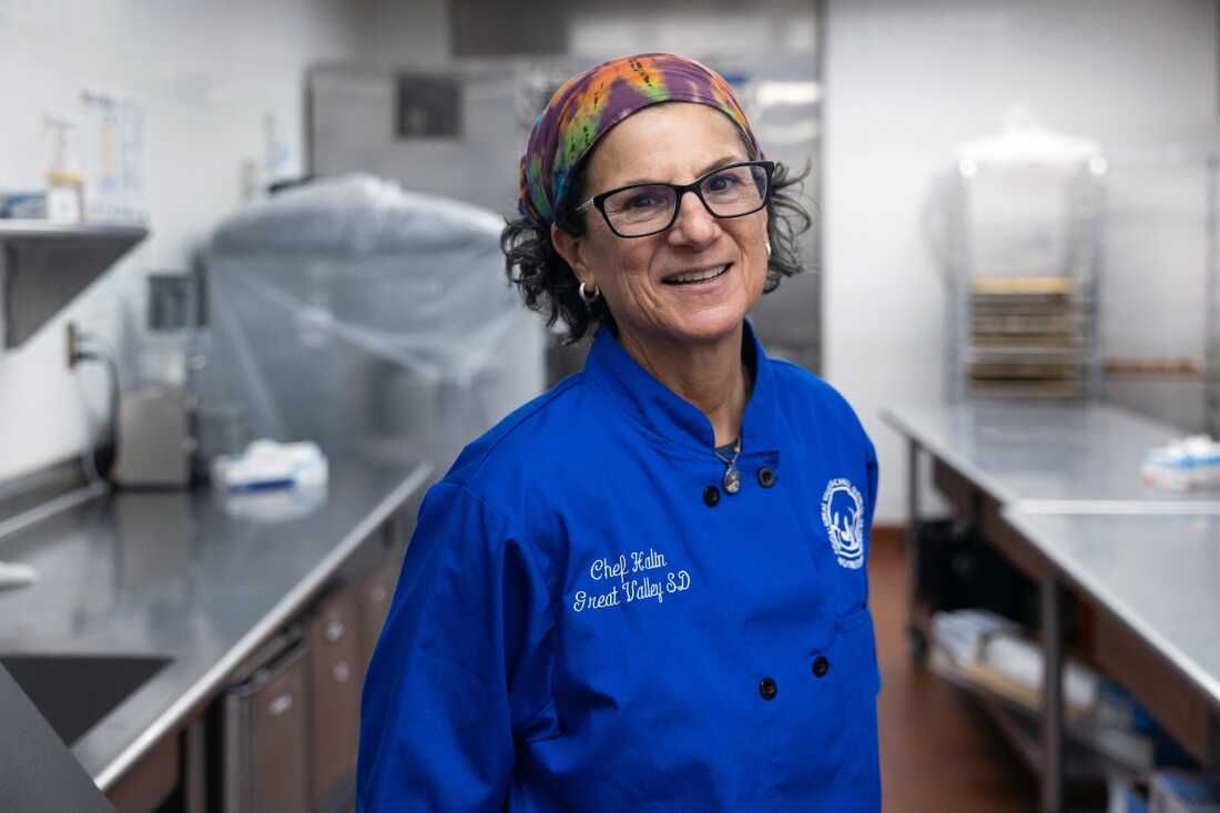 Culinary coordinator Jenifer Halin stands in the kitchen of a school cafeteria, with silver metal food-preparation surfaces behind her. She is wearing a blue chef's coat and has a multicolored bandanna on her head.