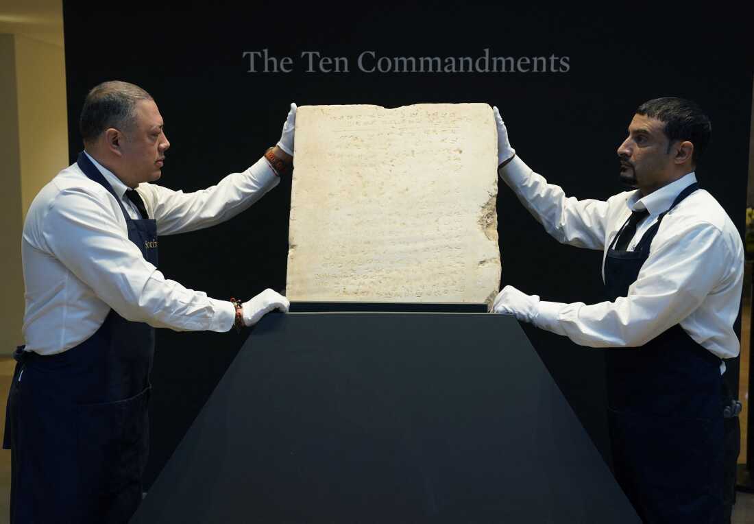 Two Sotheby's workers wearing white gloves hold a stone tablet of the Ten Commandments.