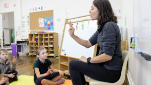 A woman is seated at the front of a classroom with children seated on colorful interlocking-mats on the floor in front of her.