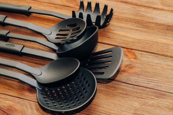 In this photo, black plastic kitchen utensils rest on a wooden surface. The items include a large serving spoon, a ladle and a slotted spatula.