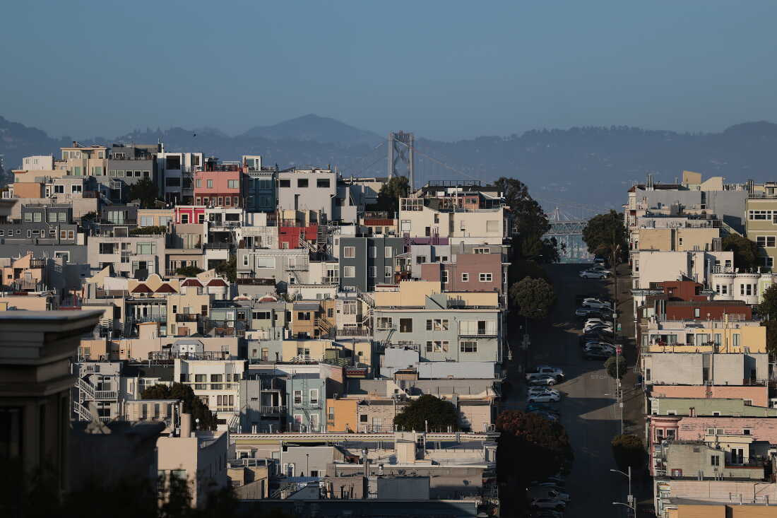 This photo taken at a distance shows houses and buildings in San Francisco's North Beach neighborhood. The structures are mainly various pastel colors such as pink, light blue and butter yellow. Mountains rise in the background.