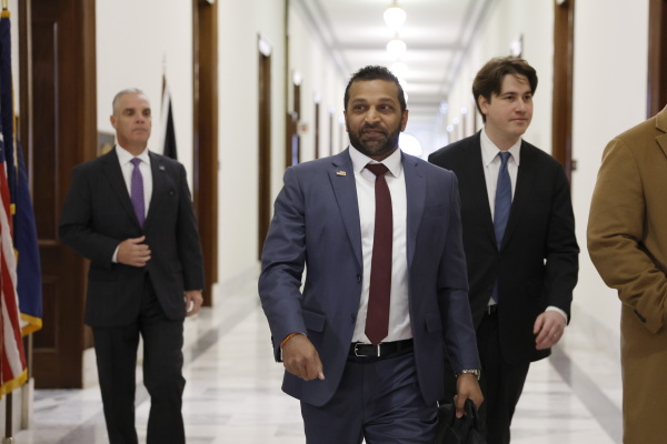 In this photo, Kash Patel is walking down a hallway. He's wearing a blue suit, and two other men in suits are walking behind him.