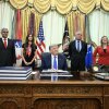 President Donald Trump sits at the Resolute Desk with several people behind him.